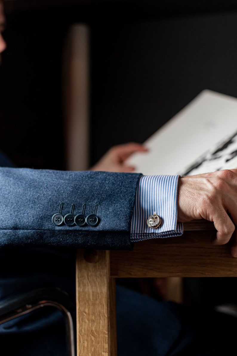 May include: A close-up of a blue blazer sleeve with four black buttons and a white and blue striped shirt cuff with a silver cufflink. The cufflink has a detailed design.