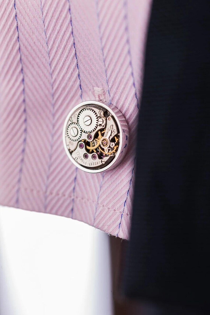 May include: A silver cufflink with a round, clear face showing the inner workings of a watch mechanism. The cufflink is on a pink and white striped fabric.