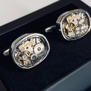 Può includere: Un paio di gemelli d'argento con un meccanismo di orologio visibile. I gemelli sono in una scatola nera.