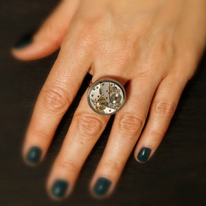 May include: A silver ring with a round, clear glass face that reveals a working clock mechanism. The ring is worn on a finger with dark green nail polish.