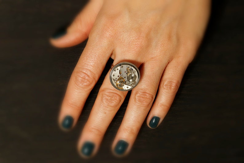 May include: A silver ring with a round, clear glass face that reveals a working clock mechanism. The ring is worn on a finger with dark green nail polish.