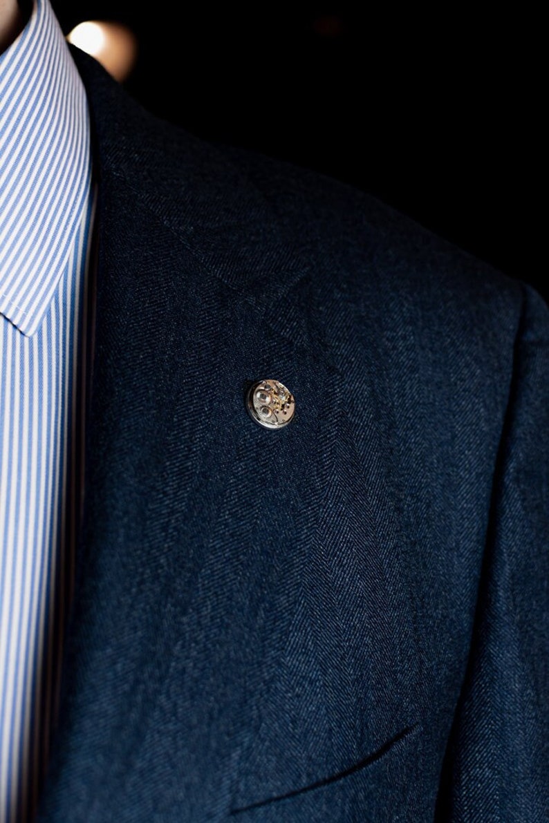 Pu&ograve; includere: Un blazer blu scuro con una spilla argentata con un design circolare con dettagli intricati.