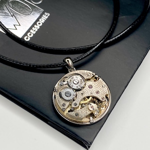 May include: A silver pendant necklace with a watch mechanism design. The round pendant displays intricate gears, jewels, and watch components. The necklace has a black leather cord and is on a black surface.