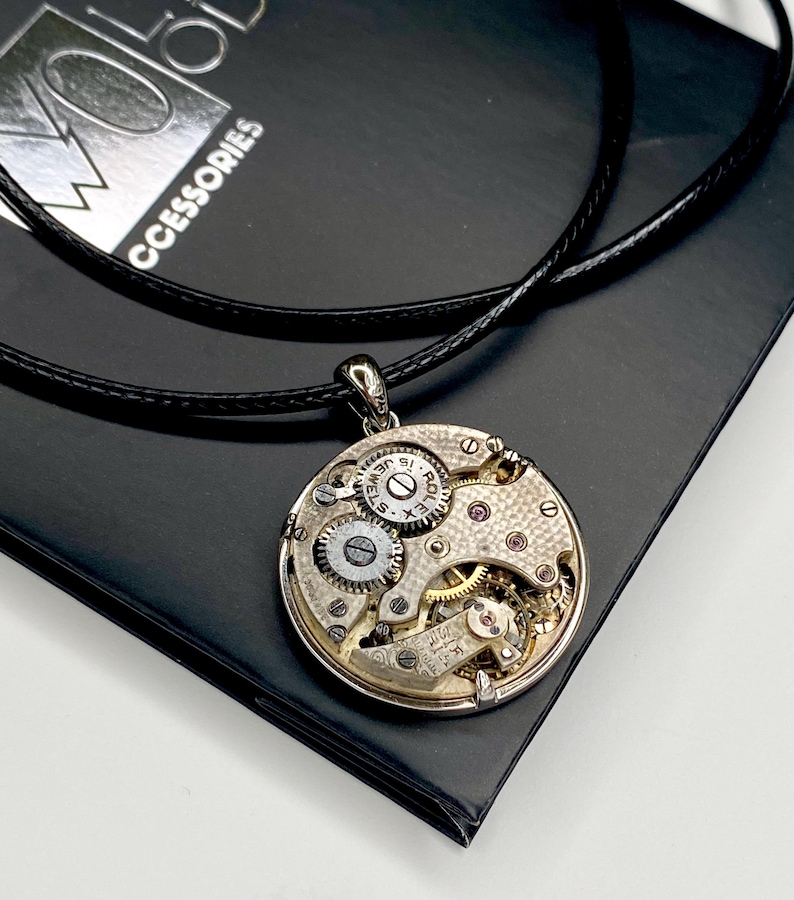 May include: A silver pendant necklace with a watch mechanism design. The round pendant displays intricate gears, jewels, and watch components. The necklace has a black leather cord and is on a black surface.