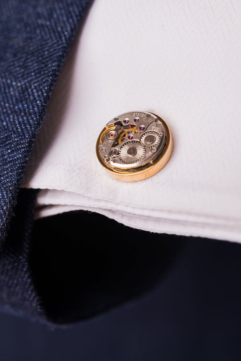 May include: A gold cufflink with a silver watch mechanism inside. The cufflink is on a white fabric surface.