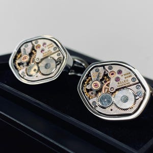 Puede incluir: Un par de gemelos plateados con mecanismos de reloj expuestos. Las piezas del reloj incluyen engranajes, joyas y el texto "ROLEX PRECISION" y "17 RUBIS SWISS". Los gemelos se muestran sobre una superficie negra.