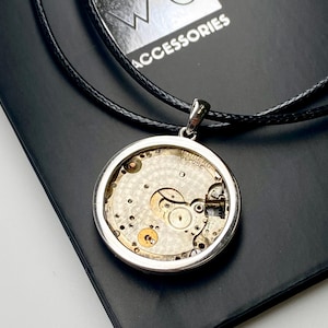 May include: A silver pendant necklace displaying the internal workings of a watch. The circular pendant reveals the gears and springs, suspended on a black cord. The word "ACCESSORIES" is visible in the background.