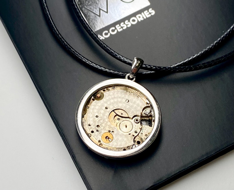May include: A silver pendant necklace displaying the internal workings of a watch. The circular pendant reveals the gears and springs, suspended on a black cord. The word "ACCESSORIES" is visible in the background.