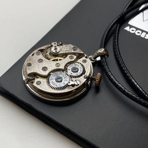 May include: A silver pendant necklace crafted from a repurposed watch mechanism. The intricate internal workings of the watch are displayed, including gears and jewels. The pendant is suspended from a black cord, set against a dark background.