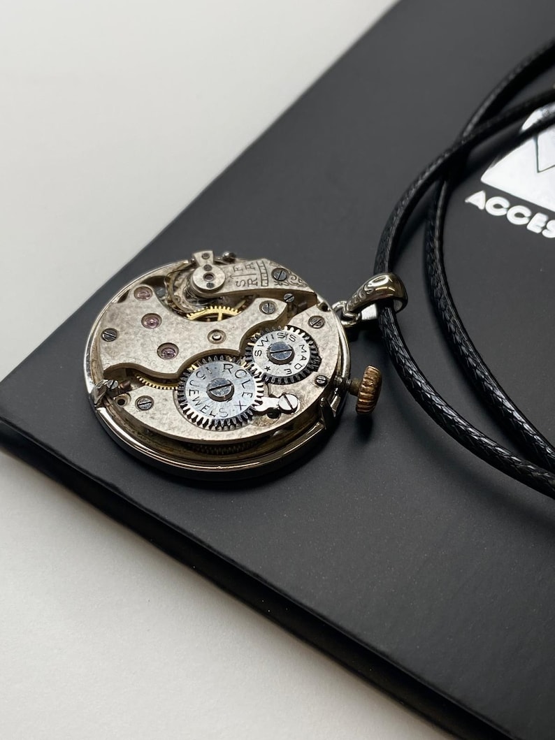 May include: A silver pendant necklace crafted from a repurposed watch mechanism. The intricate internal workings of the watch are displayed, including gears and jewels. The pendant is suspended from a black cord, set against a dark background.