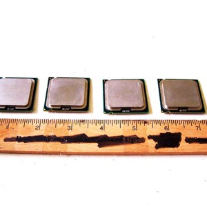 Op de afbeelding: Vier vierkante computerprocessors met een zilveren bovenkant en groene randen worden getoond. Een houten liniaal met zwarte markeringen is eronder geplaatst, gemeten in centimeters. De processors zijn in een rij gerangschikt.