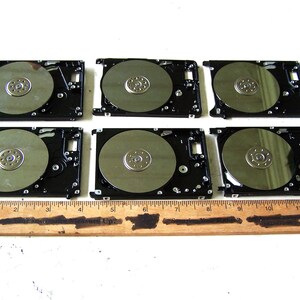 May include: Six disassembled hard drive components with exposed circular silver platters and black metal frames, arranged in two rows of three, positioned behind a wooden ruler that measures up to 30 cm in length on a white surface.