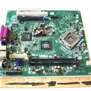May include: A green Dell AT3422 computer motherboard with various electronic components, including chips, connectors, and slots. The board has a rectangular shape and features multiple colours, including green, purple, and white. A wooden ruler is at the bottom.