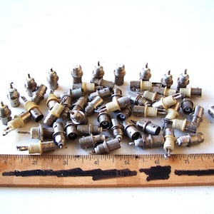 May include: A pile of vintage spark plugs, made from metal and ceramic. The plugs are scattered on a white surface, with a wooden ruler for scale. The plugs appear to be used.
