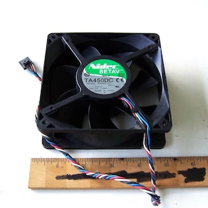 Puede incluir: Un ventilador de ordenador negro con una etiqueta verde y blanca que dice "Nidec BETAV" y "TA450DC". El ventilador tiene aspas negras y un marco cuadrado. Una regla de madera está colocada debajo del ventilador. El ventilador tiene un cable multicolor.