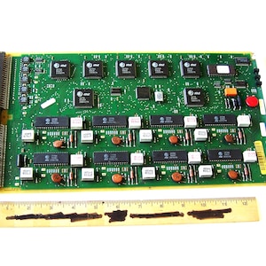 May include: A green circuit board with numerous black chips, white components, and orange capacitors. The board has a gold edge and a black connector on the left side. A wooden ruler is at the bottom, and the board has the AT&T logo.