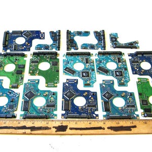 May include: A collection of fourteen assorted blue and green printed circuit boards of varying shapes and sizes, each featuring integrated chips and connectors, arranged in rows above a wooden ruler measuring twelve inches in length.