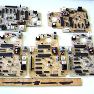 Puede incluir: Varias placas de circuito beige con diversos componentes electrónicos, incluyendo chips y conectores. Una regla de madera con marcas en centímetros se encuentra en la parte inferior. Las placas tienen formas y tamaños variados.