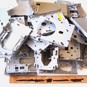 May include: A collection of silver metal computer parts, such as brackets and plates, in various shapes and sizes. A wooden ruler with markings is at the bottom of the pile, showing measurements.