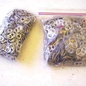 May include: Two clear plastic bags filled with a large quantity of silver-colored aluminum soda can pull tabs. The bag on the left is sealed, while the bag on the right features a red and white zipper closure at the top containing mixed tabs.