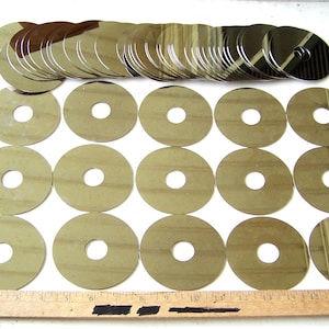May include: A collection of reflective, circular discs with a central hole, arranged on a white surface. The discs are a light, metallic color, and a wooden ruler is at the bottom. The discs are approximately 2 inches in diameter.