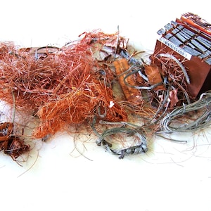 May include: A collection of scrap metal, including tangled copper wires, a brown rectangular component, and various other metallic pieces. The copper wires are a vibrant orange color, contrasting with the silver and brown tones of the other components.