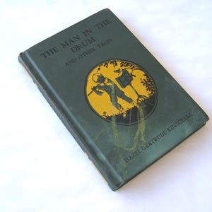 May include: A green book titled "The Man in the Drum and Other Tales" by Hazel Gertrude Kinscella. The cover features a black and white illustration of a boy playing a drum in a yellow circle.