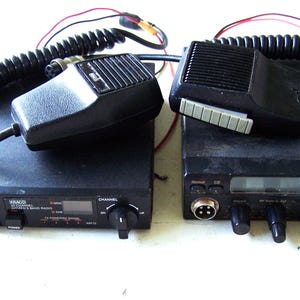 May include: Two black CB radios with coiled microphones. The left unit is a Kraco 40-channel citizen's band radio with volume, squelch, and channel knobs. The right unit is a Cobra 19 DX IV radio with a digital display, knobs, and a microphone port.