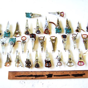 May include: A collection of thirty-three silver-toned metal hard drive actuator arms with various coloured components, arranged in rows above a wooden ruler measuring 30.5 cm in length, showing the scale of these electronic components.