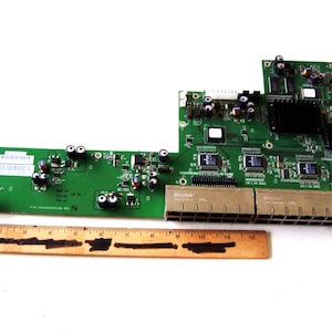 May include: A green circuit board with various electronic components, including chips, connectors, and a black heat sink. A wooden ruler is placed below the board. The board appears to be a network device.
