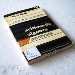 Op de afbeelding: Een paperbackboek getiteld Elementary Mathematics from an Advanced Standpoint door Felix Klein, met een zwart-crèmekleurige omslag en de woorden arithmetic, algebra en analysis in het wit gedrukt. Een bibliotheeklabel toont de code QA 39 K54 1948a.