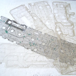 May include: A collection of translucent, flexible membrane circuit sheets featuring intricate conductive pathways and circular contact points. One sheet displays a prominent black grid pattern overlay with a metallic connector component attached.
