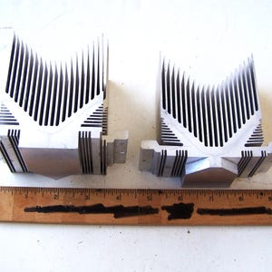 May include: Two silver-colored heat sinks with a V-shaped design and vertical fins. The heat sinks are made of metal and have a rectangular base. A wooden ruler with inch markings is in the foreground.