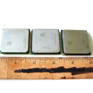 Op de afbeelding: Drie vierkante zilverkleurige computerprocessors met groene printplaatvoeten, geplaatst boven een houten liniaal die laat zien dat de processors elk ongeveer 3,8 cm breed zijn. De metalen oppervlakken zijn voorzien van geëtste tekst en QR-codes.