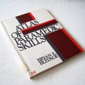 May include: A hardcover book titled "Atlas of Paramedic Skills" by Bryan E. Bledsoe. The book has a white cover with black, red, and gray text and design elements. The book is slightly angled, showing the spine and a small label.