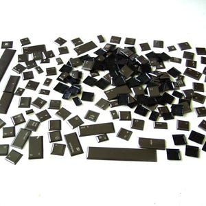 Op de afbeelding: Een grote verzameling zwarte plastic toetsenbordtoetsen, verspreid over een wit oppervlak. De toetsen hebben verschillende letters, cijfers en symbolen erop gedrukt.