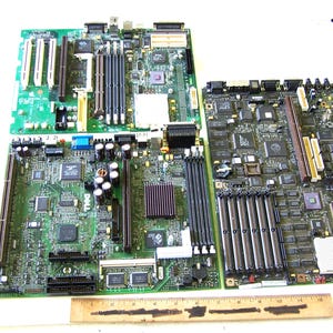 May include: Three rectangular computer motherboards featuring various integrated circuits, memory slots, and expansion ports. A wooden ruler is positioned at the bottom, showing a scale of 12 inches to provide a clear sense of the physical size.
