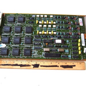 May include: A green circuit board with numerous integrated circuits, resistors, and other electronic components. A wooden ruler is placed below the board. The board has a variety of colors, including green, black, yellow, and white.