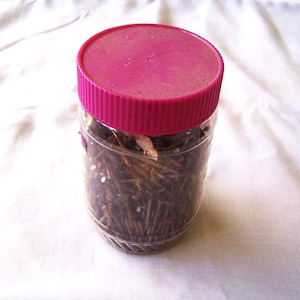 Op de afbeelding: Een doorzichtige cilindervormige plastic pot gevuld met talloze donkerbruine metalen spijkers. De pot heeft een felroze schroefdeksel met een geribbelde rand. De container is transparant, waardoor de metalen inhoud goed zichtbaar is.