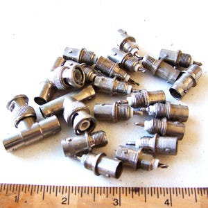 May include: A collection of silver BNC connectors, in various shapes and sizes, scattered on a white surface. Some connectors are cylindrical, others angled. A wooden ruler with centimetre markings is visible at the bottom.