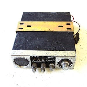 Puede incluir: Un transceptor CB Regency CR-185 negro con un panel frontal plateado que incluye un medidor de señal redondo, cuatro perillas de control y tres interruptores. Un soporte de montaje de metal dorado está fijado sobre la unidad.