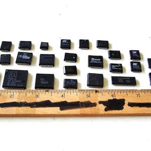 May include: A collection of black integrated circuit chips of various sizes, arranged on a white surface. A wooden ruler with inch markings is placed at the bottom, providing scale. The chips have text and markings.