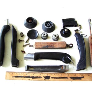 May include: A collection of assorted kitchen hardware including black plastic handles, metal knobs, screws, and wooden tool components arranged above a 12-inch wooden ruler. The items consist of various replacement parts for cookware and utensils.