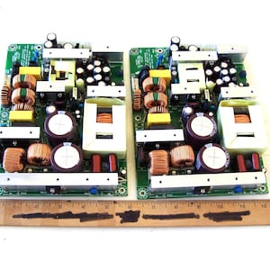 May include: Two electronic circuit boards with various components, including copper coils, capacitors, and transformers. The boards are green with silver and yellow elements. A wooden ruler is placed below the boards.