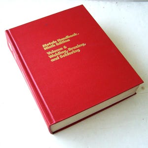 May include: A red hardcover book titled "Metals Handbook, Ninth Edition, Volume 6, Welding, Brazing, and Soldering" in gold lettering. The book has a clean, simple design.
