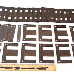 May include: A collection of rusty metal components, including rectangular strips with square cutouts and "E" shaped pieces with small holes. A wooden ruler with black markings is at the bottom.