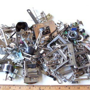 May include: A pile of various silver-colored sewing machine parts, including presser feet, bobbins, and other metal components. A wooden ruler with inch markings is at the bottom of the image.