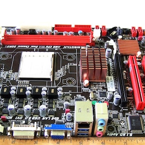 May include: A black motherboard with red memory slots and a copper-colored heatsink. A wooden ruler is positioned along the bottom edge, showing the board measures approximately 9.5 inches in width. It features various ports and capacitors.