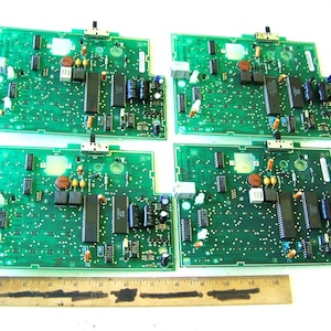 May include: Four rectangular green printed circuit boards featuring various electronic components, including integrated circuits, capacitors, and resistors. A wooden ruler is positioned below the boards, showing a length of approximately 12 inches.