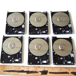 Könnte beinhalten: Sechs Festplatten mit silbernen Tellern und schwarzen Gehäusen sind auf einer weißen Oberfläche angeordnet. Ein Holzlineal mit Zollmarkierungen befindet sich am unteren Bildrand und dient als Maßstab.